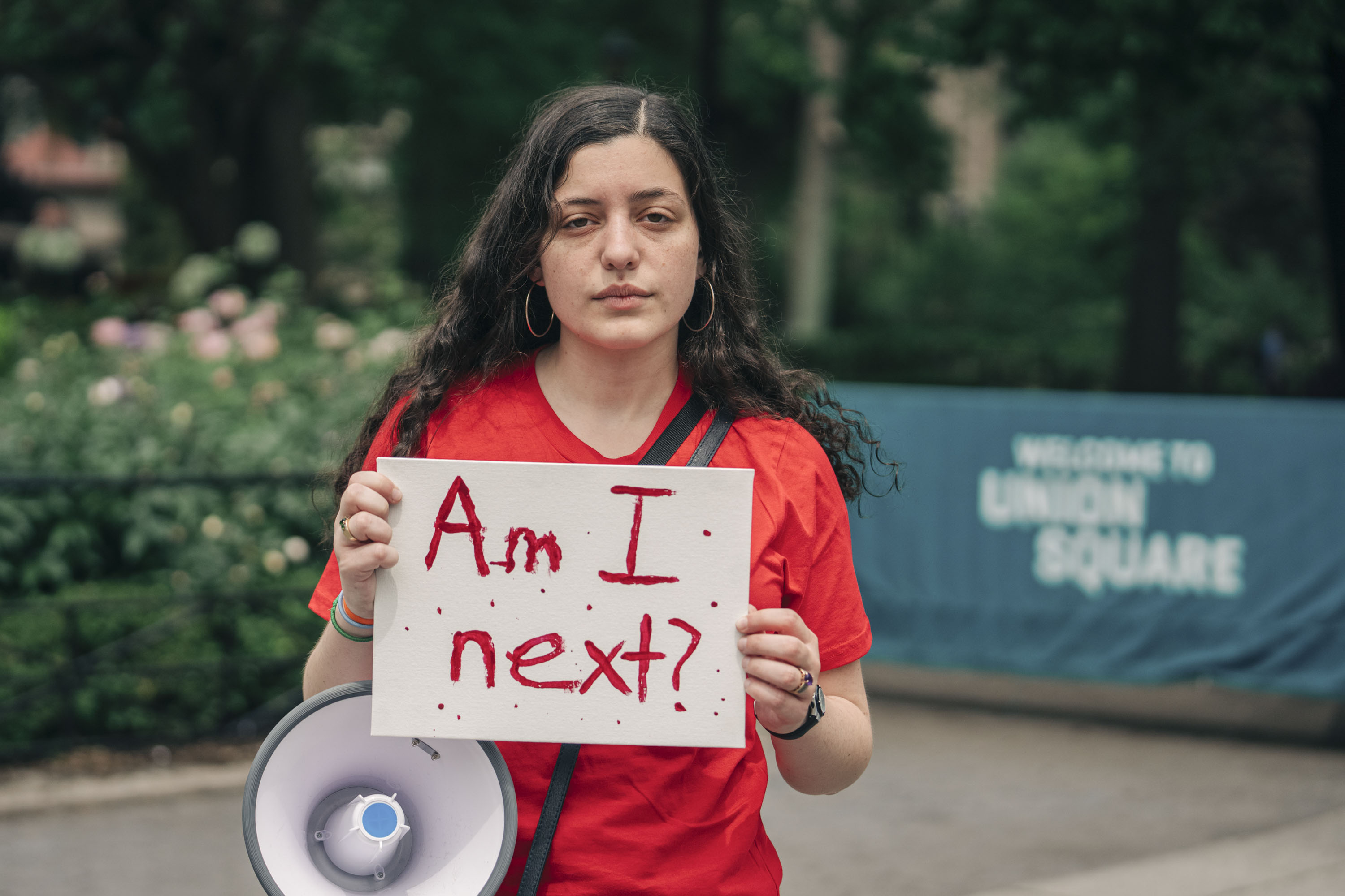 In a public park, a teenager/young adult in a red shirt is carrying a megaphone and holding a protest sign that says "Am I next?" with a somber expression.