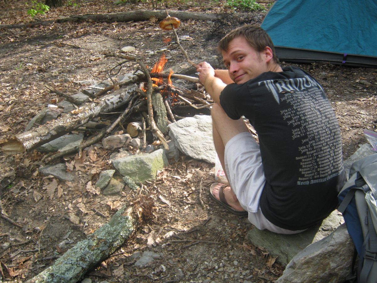 Photo of Dorothy Paugh's late son, Peter, smiling and looking over his shoulder while roasting a bagel at a campfire.